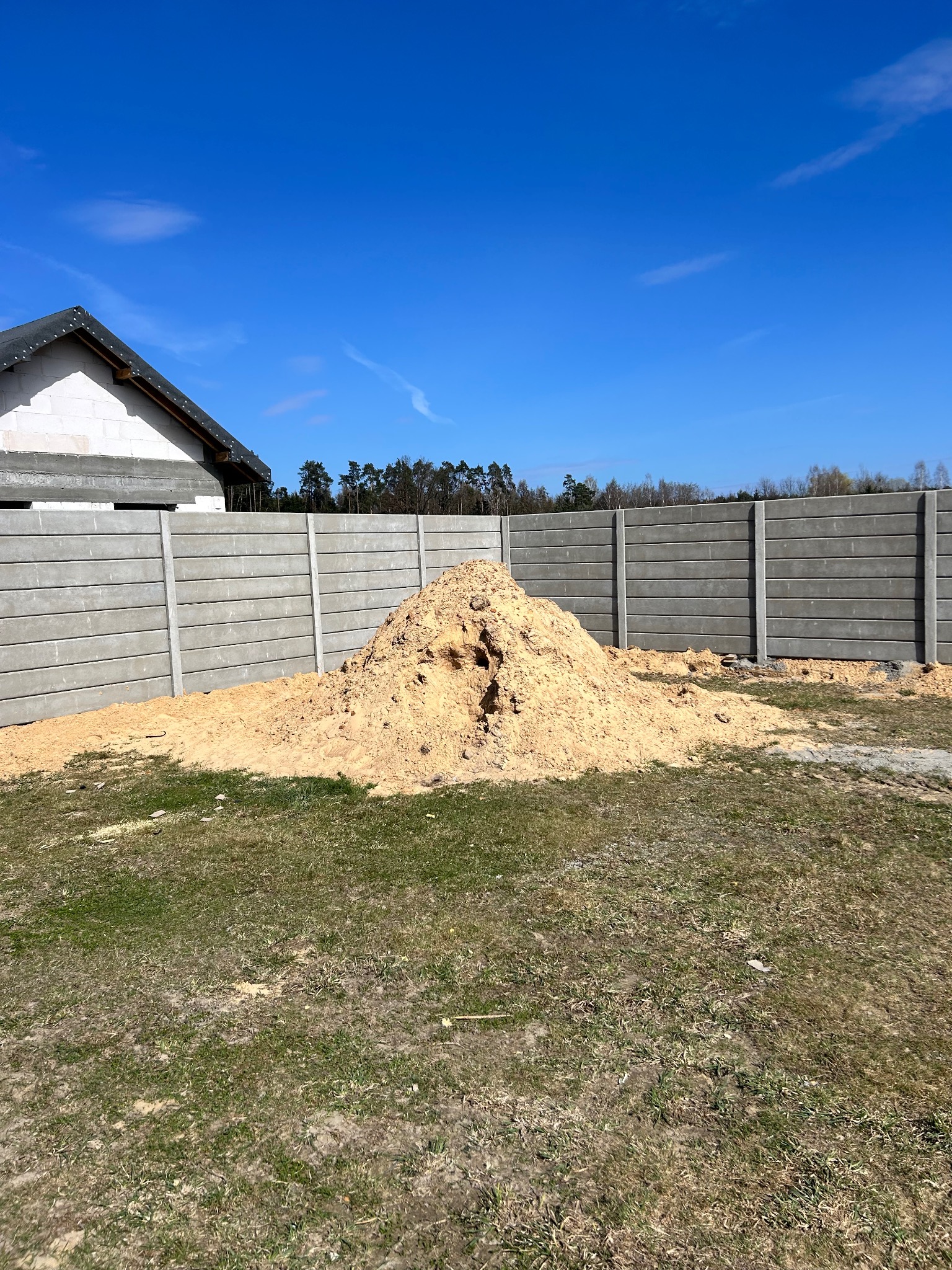 Szary, prefabrykowany płot betonowy na trawiastym terenie budowy, z hałdą piasku i fragmentem domu w tle, w słoneczny dzień.