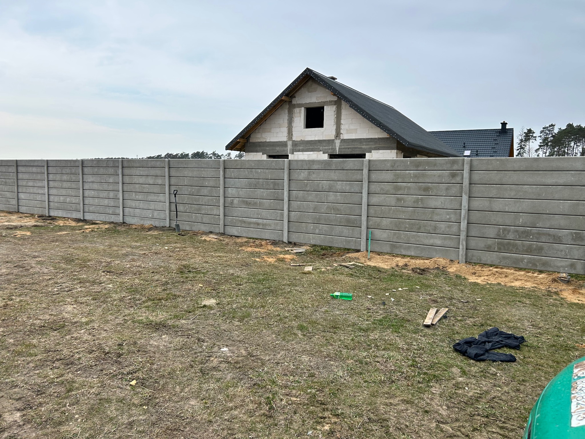 Szary, prefabrykowany płot betonowy z widoczną szpadą, na tle budowy domu jednorodzinnego w stanie surowym zamkniętym, w pochmurny dzień.