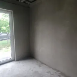 Surowe wnętrze pomieszczenia po nałożeniu tynku maszynowego na ściany i wylewki na podłogę, widoczne drewniane elementy konstrukcyjne sufitu i okno z widokiem na zieleń.