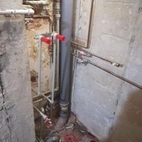 Niedokończona instalacja wodno-kanalizacyjna w starej łazience z surowymi ścianami, widoczne rury PCV i metalowe, zawory z czerwonymi uchwytami, brak płytek.