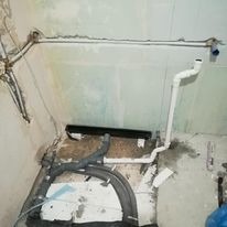 Surowy stan łazienki w trakcie prac: widoczne rury kanalizacyjne i wodne, częściowo zakryte płytą kartonowo-gipsową, zarys odpływu liniowego w podłodze, ściany w stanie deweloperskim z widocznymi...