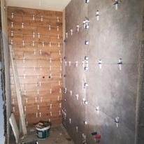 Ściana łazienki w trakcie układania płytek imitujących drewno, widoczne krzyżyki dystansowe i poziomica oparta o ścianę. Druga ściana wyłożona szarymi płytkami, również w trakcie montażu. Wiadro...