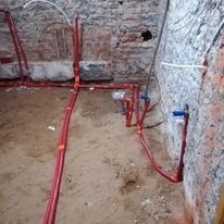 Czerwone rury instalacji wodnej ułożone na piasku, widoczne fragmenty ceglanej ściany w trakcie remontu, nieotynkowane ściany, brak wykończenia.