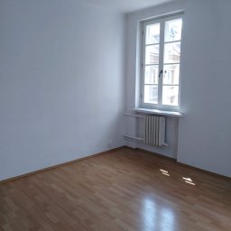 manius-bud Mariusz Witan - Pusty pokój z białymi ścianami, drewnianą podłogą i oknem z białymi ramami, grzejnik pod oknem, widoczne ślady po niedawno zakończonych pracach.