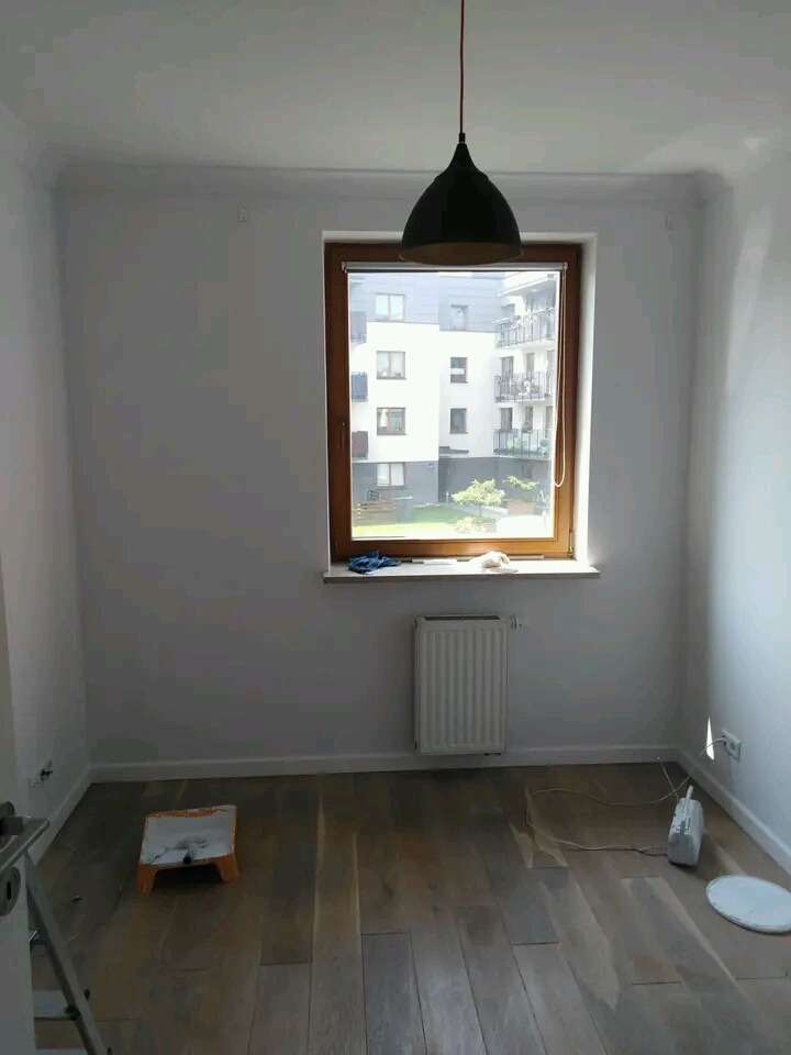 Pokój po odświeżeniu koloru ścian na biało, widoczny grzejnik, okno z drewnianą ramą i lampa wisząca, na podłodze drewniane panele i akcesoria malarskie.