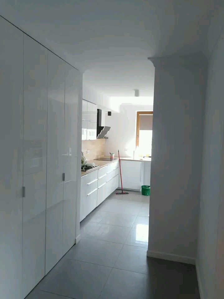 Wnętrze po malowaniu: Biała, minimalistyczna kuchnia z błyszczącymi frontami szafek, szarymi płytkami na podłodze i czerwonym mopem opartym o ścianę, widok z korytarza.