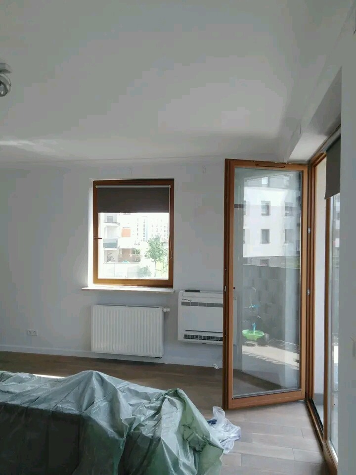 Pokój przed pracami wykończeniowymi: zabezpieczone meble folią malarską, świeżo pomalowane białe ściany, okno z brązową ramą i roletą, grzejnik pod oknem.