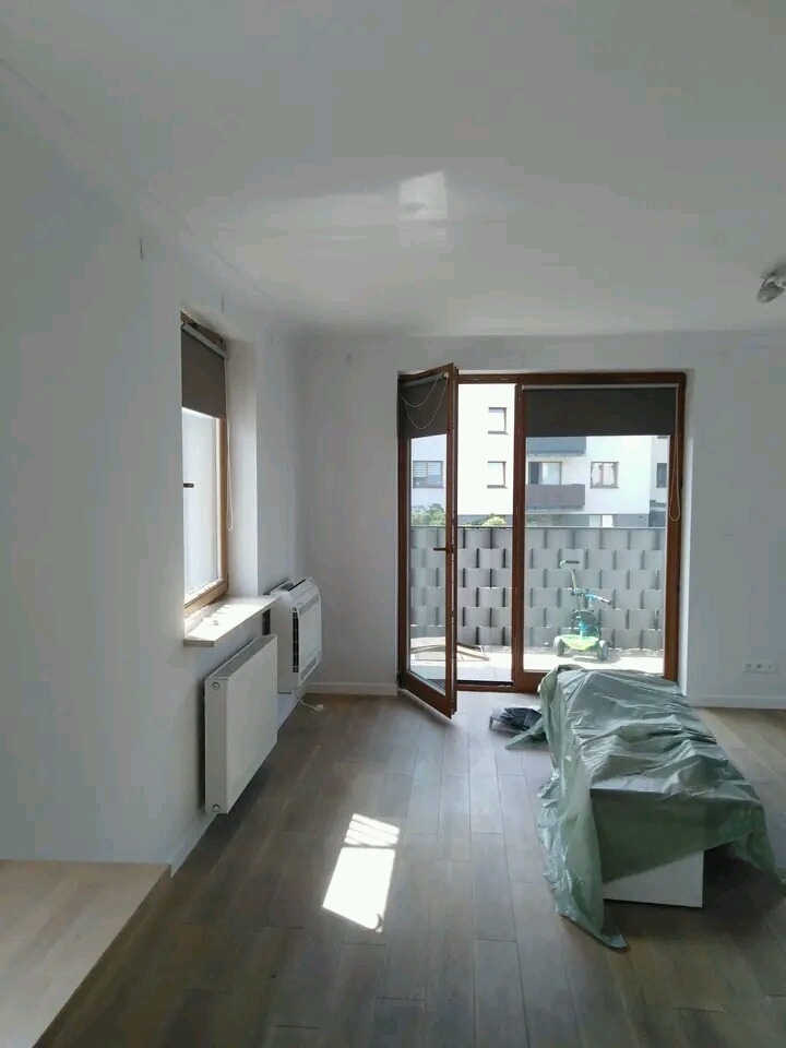 Wnętrze mieszkania w Warszawie po świeżym pomalowaniu ścian na biało, z widocznymi grzejnikami, drewnianą podłogą i balkonem z widokiem na zewnątrz.