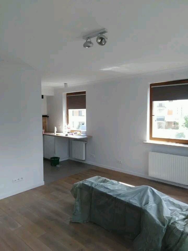 Jasne wnętrze po remoncie, świeżo pomalowane ściany na biało, zabezpieczone meble folią ochronną, parkiet na podłodze, widoczne okna z brązowymi ramami i grzejniki.