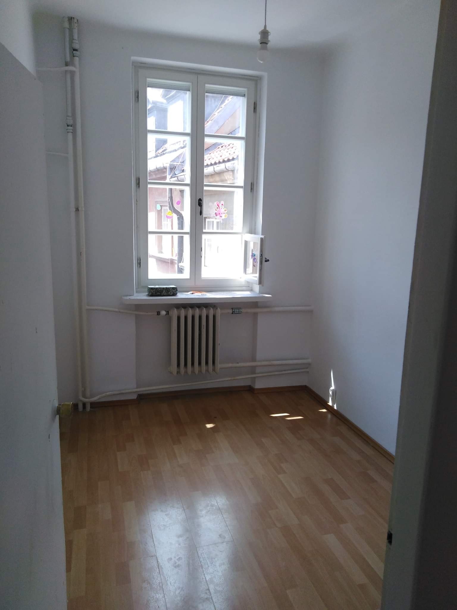 Pusty pokój z białymi ścianami, parkietem i grzejnikiem pod oknem, widoczne rury grzewcze przy ścianie, pojedyncza żarówka na suficie.
