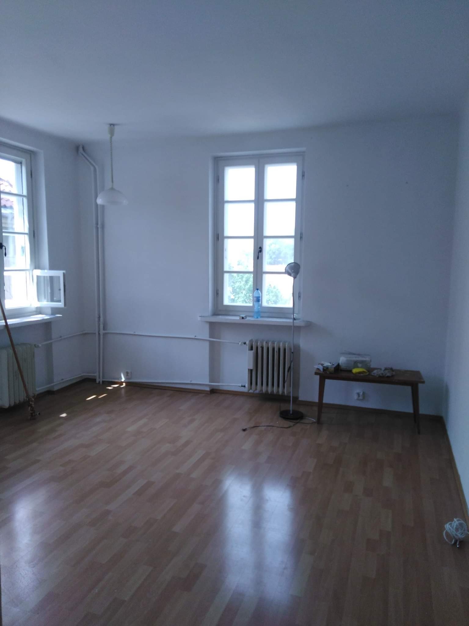 Puste, jasne pomieszczenie z parkietem, grzejnikami pod oknami i stołem z akcesoriami malarskimi, przygotowane do odświeżenia ścian.