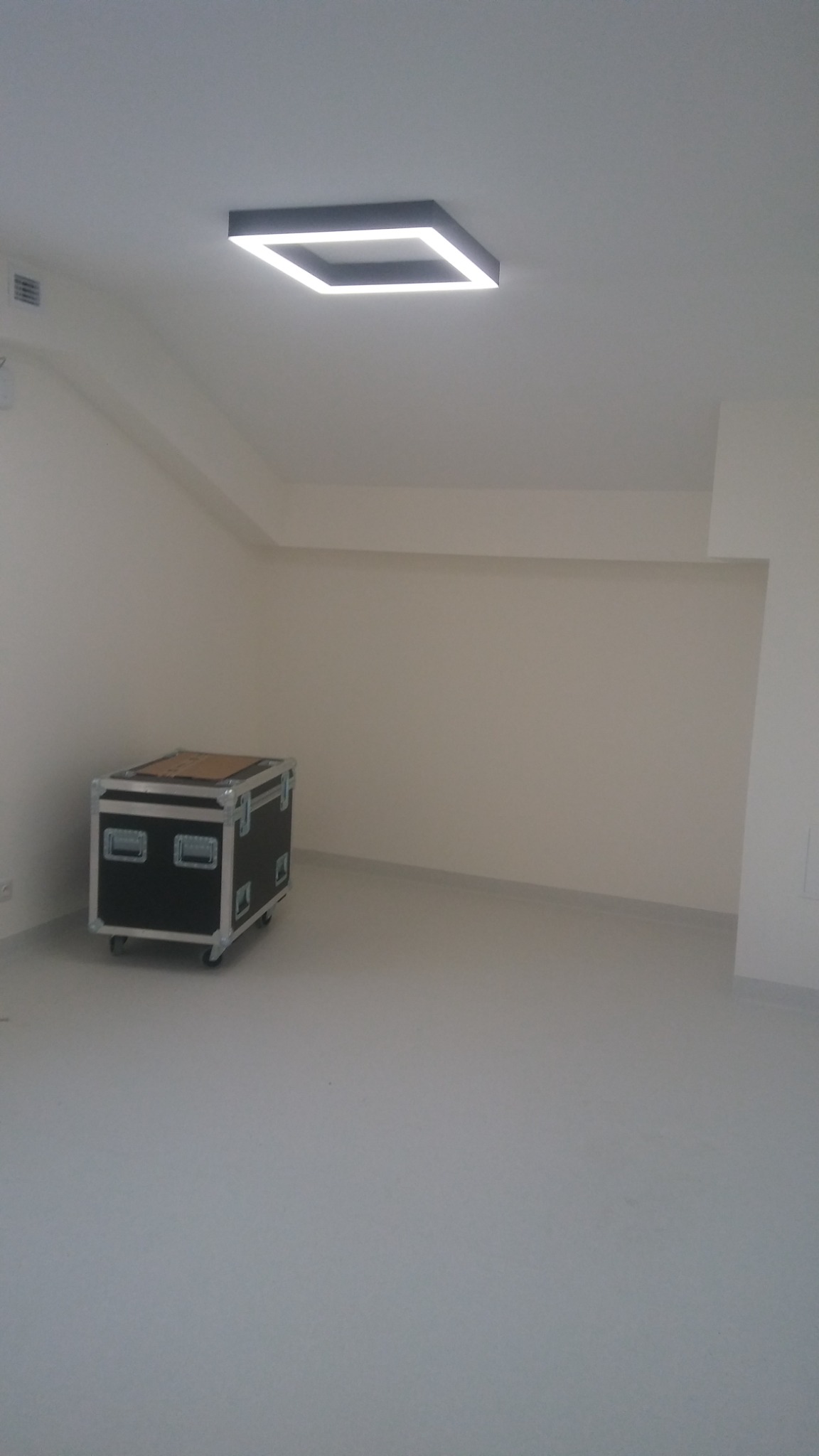 Puste, świeżo pomalowane pomieszczenie z białymi ścianami i szarą podłogą, oświetlone nowoczesną, kwadratową lampą LED w suficie, z czarną skrzynią transportową na kółkach po lewej stronie.
