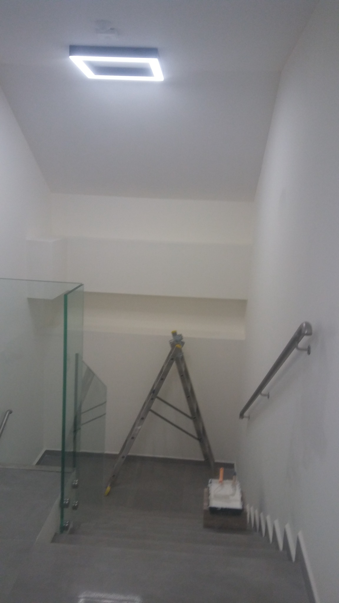 Klatka schodowa w trakcie odświeżania, widoczna drabina aluminiowa, wałek malarski z kuwetą na schodach oraz szklana balustrada.
