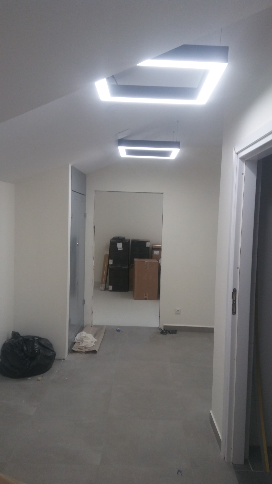 Wnętrze w trakcie remontu: świeżo pomalowane ściany w odcieniach bieli, szara podłoga, nowoczesne kwadratowe lampy sufitowe, widoczne kartony i worki z odpadami budowlanymi.