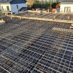 Zbrojenie płyty fundamentowej domu jednorodzinnego z wystającymi prętami pionowymi, widok z góry na tle budynków w zabudowie szeregowej.