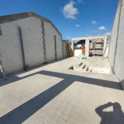 Wnętrze domu w trakcie budowy z białego bloczka, widoczne ściany, betonowa podłoga z otworem na schody, podpory i rury, w tle rusztowanie i budynki.