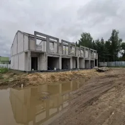 Wznoszenie szeregowca z betonu komórkowego, widoczne niezadaszone surowe ściany działowe i słupy nośne, teren budowy z kałużami i śladami opon.