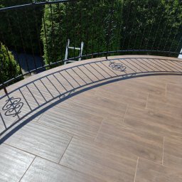 Usługi Ogólnobudowlane Dariusz Kunicki - Balkon z brązowymi płytkami imitującymi drewno i czarną, metalową balustradą z ozdobnymi elementami, na tle zielonych krzewów.