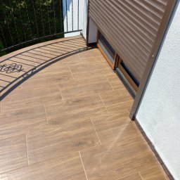 Usługi Ogólnobudowlane Dariusz Kunicki - Wykończony balkon z brązowymi płytkami imitującymi drewno, częściowo opuszczona roleta zewnętrzna i czarna metalowa balustrada.