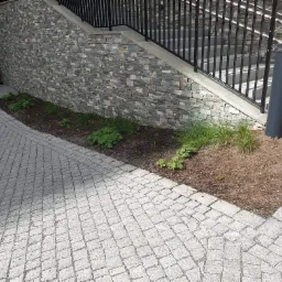 Kamienna ściana z schodami i metalową balustradą, obok kostka brukowa ułożona w romby i rabata z młodymi roślinami, oświetlona przez szarą lampę.