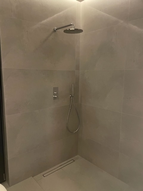 Nowoczesny prysznic typu walk-in z szarymi płytkami wielkoformatowymi, deszczownicą, odpływem liniowym i minimalistyczną baterią podtynkową.