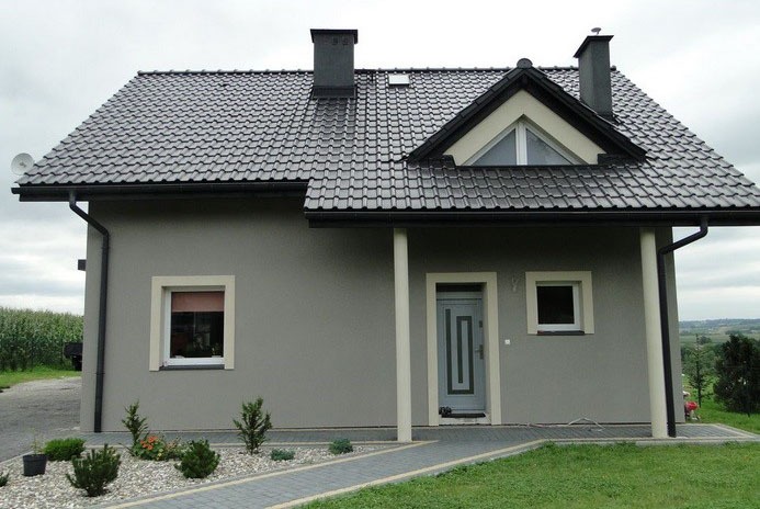 Nowoczesny dom jednorodzinny z szarą elewacją, ciemną dachówką i jasnymi obramowaniami okien, otoczony niską roślinnością i ścieżką z kostki brukowej.