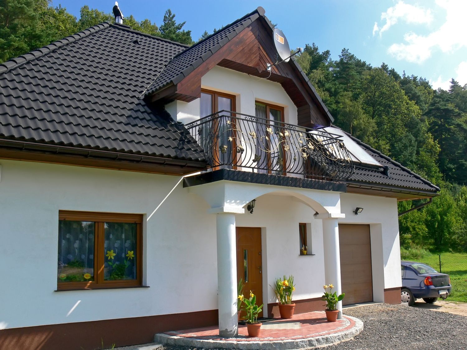 Biały dom jednorodzinny z czarnym dachem i balkonem z kutą balustradą, widok z zewnątrz, z elementami dekoracyjnymi i zadbanym otoczeniem, w tle las.