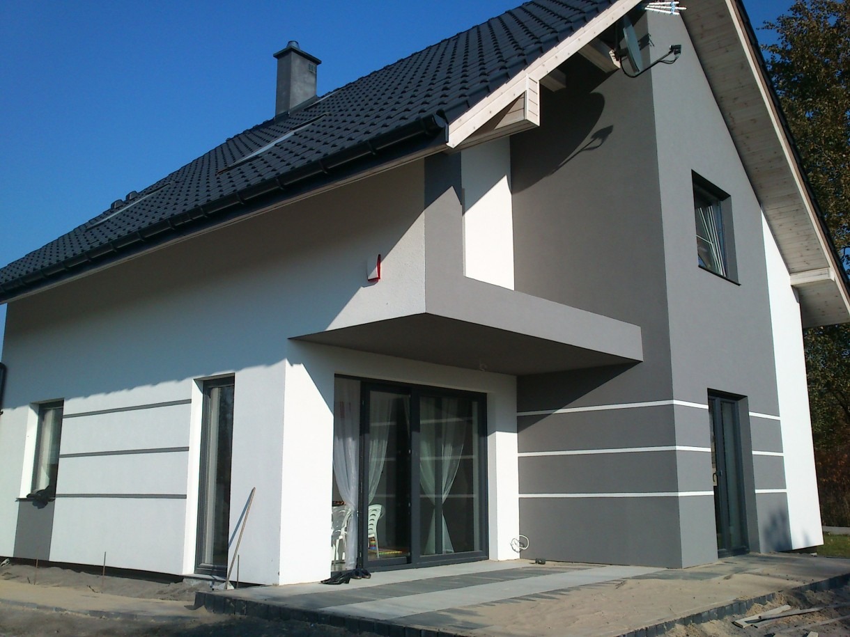 Nowoczesny dom jednorodzinny z szarą i białą elewacją, czarnym dachem i elementami dekoracyjnymi w postaci poziomych pasków, widoczne okna i drzwi balkonowe z zasłonami, teren wokół domu w trakcie...