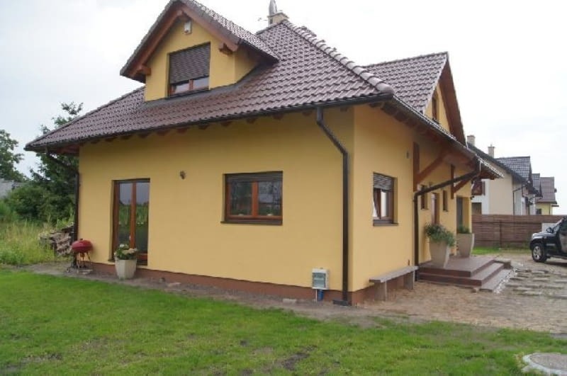 Żółty dom jednorodzinny z brązowym dachem i stolarką okienną, widok z zewnątrz na elewację i fragment ogrodu z grillem.