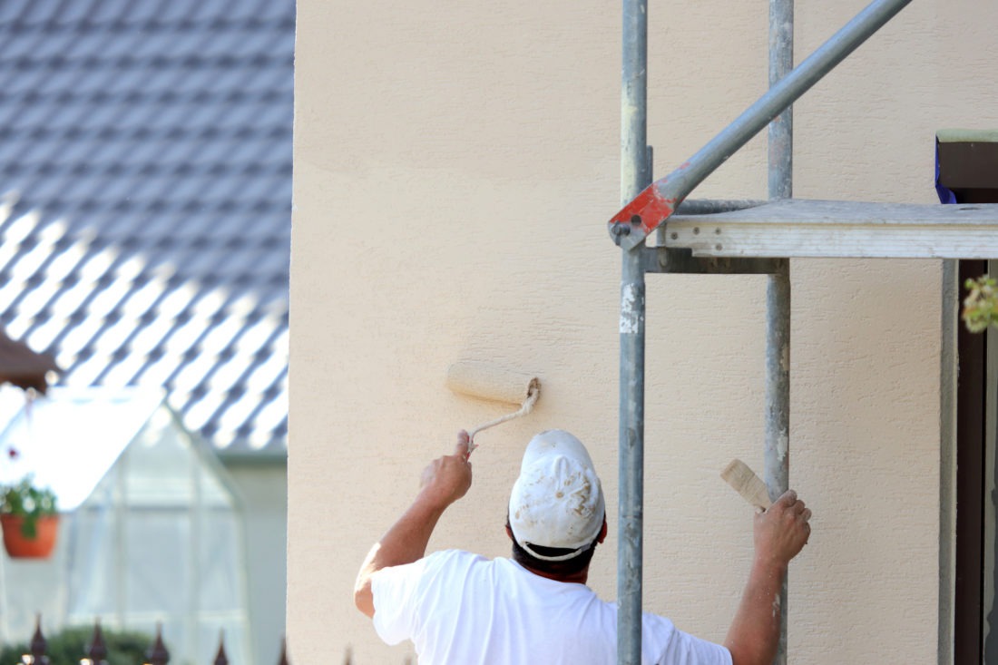 Mężczyzna w białej koszulce i czapce maluje jasną elewację budynku wałkiem, stojąc na metalowym rusztowaniu; w tle widoczny fragment dachu i szklarni.