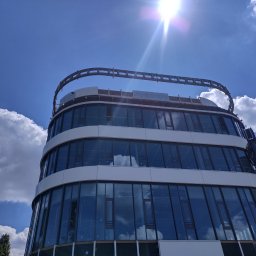 HBK BUDOWNICTWO s.c. - Nowoczesny budynek biurowy z zaokrąglonymi ścianami i szklaną fasadą, częściowo zasłonięty przez ramię dźwigu, widoczne niebo z chmurami odbijające się w szybach.