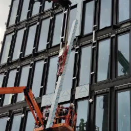 Montaż paneli elewacyjnych Alucobond na fasadzie budynku biurowego przy użyciu podnośnika koszowego i żurawia. Widoczne elementy mocujące i okna. Prace w toku.