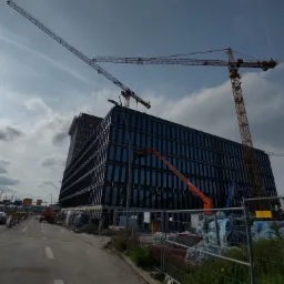 Budynek biurowy w trakcie budowy z ciemną szklaną fasadą, widoczne dwa żurawie budowlane, podnośnik koszowy, ogrodzenie placu budowy oraz ruch uliczny w tle.