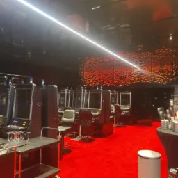 Wnętrze kasyna z rzędem automatów do gier oddzielonych pleksiglasem, czerwona wykładzina, czarne ściany z dekoracją imitującą płomienie, stolik z szklankami i wózkiem barowym.