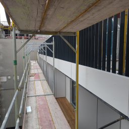 HBK BUDOWNICTWO s.c. - Widok rusztowania z drewnianymi podestami wzdłuż ściany budynku z zamontowanymi białymi panelami elewacyjnymi i czarnymi pionowymi elementami dekoracyjnymi. W tle widoczne kolejne rusztowania...