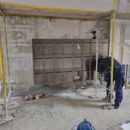 HBK BUDOWNICTWO s.c. - Montaż skrzynek pocztowych ze stali nierdzewnej na surowej ścianie wewnątrz budynku, widoczne rusztowanie i materiały budowlane, pracownik pochylony nad instalacją.