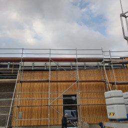 HBK BUDOWNICTWO s.c. - Montaż drewnianej elewacji z wykorzystaniem rusztowania i podnośnika, widoczny pracownik w trakcie wykonywania prac na wysokości.