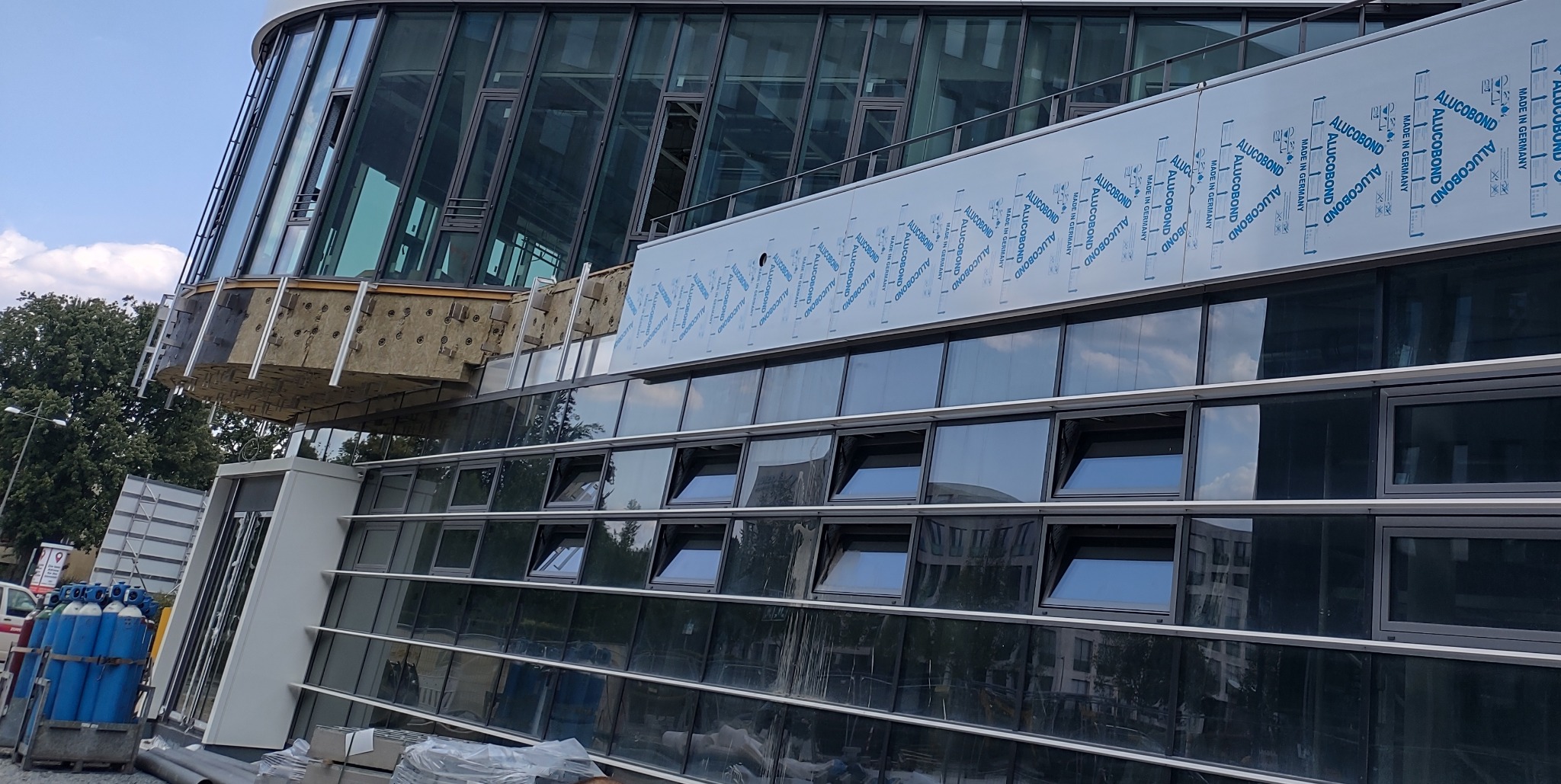 Budynek biurowy w trakcie montażu elewacji z płyt warstwowych Alucobond, widoczne otwarte okna, izolacja termiczna pod elementem konstrukcyjnym i butle z gazem na placu budowy.