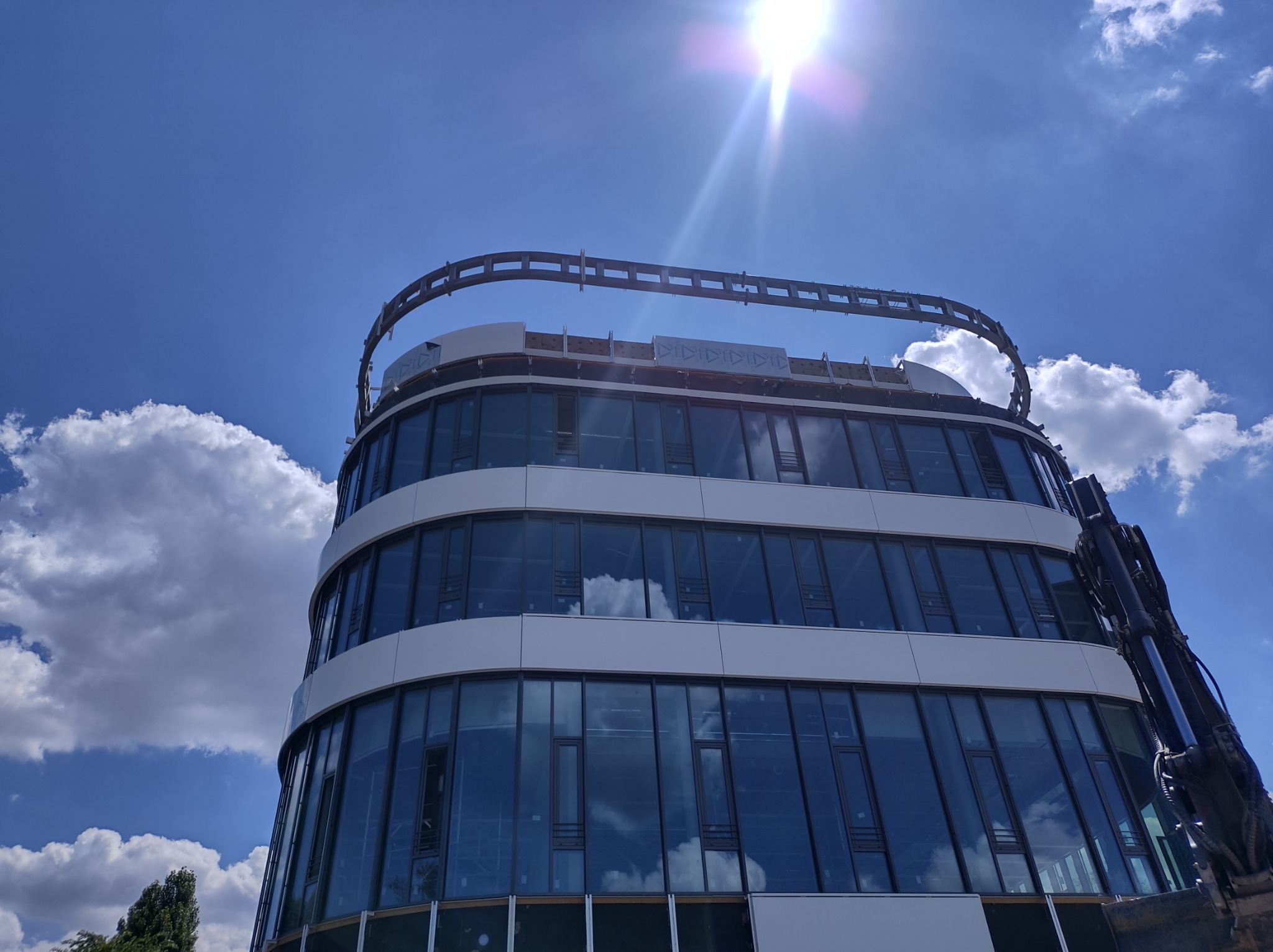 Nowoczesny budynek biurowy z zaokrąglonymi ścianami i szklaną fasadą, częściowo zasłonięty przez ramię dźwigu, widoczne niebo z chmurami odbijające się w szybach.