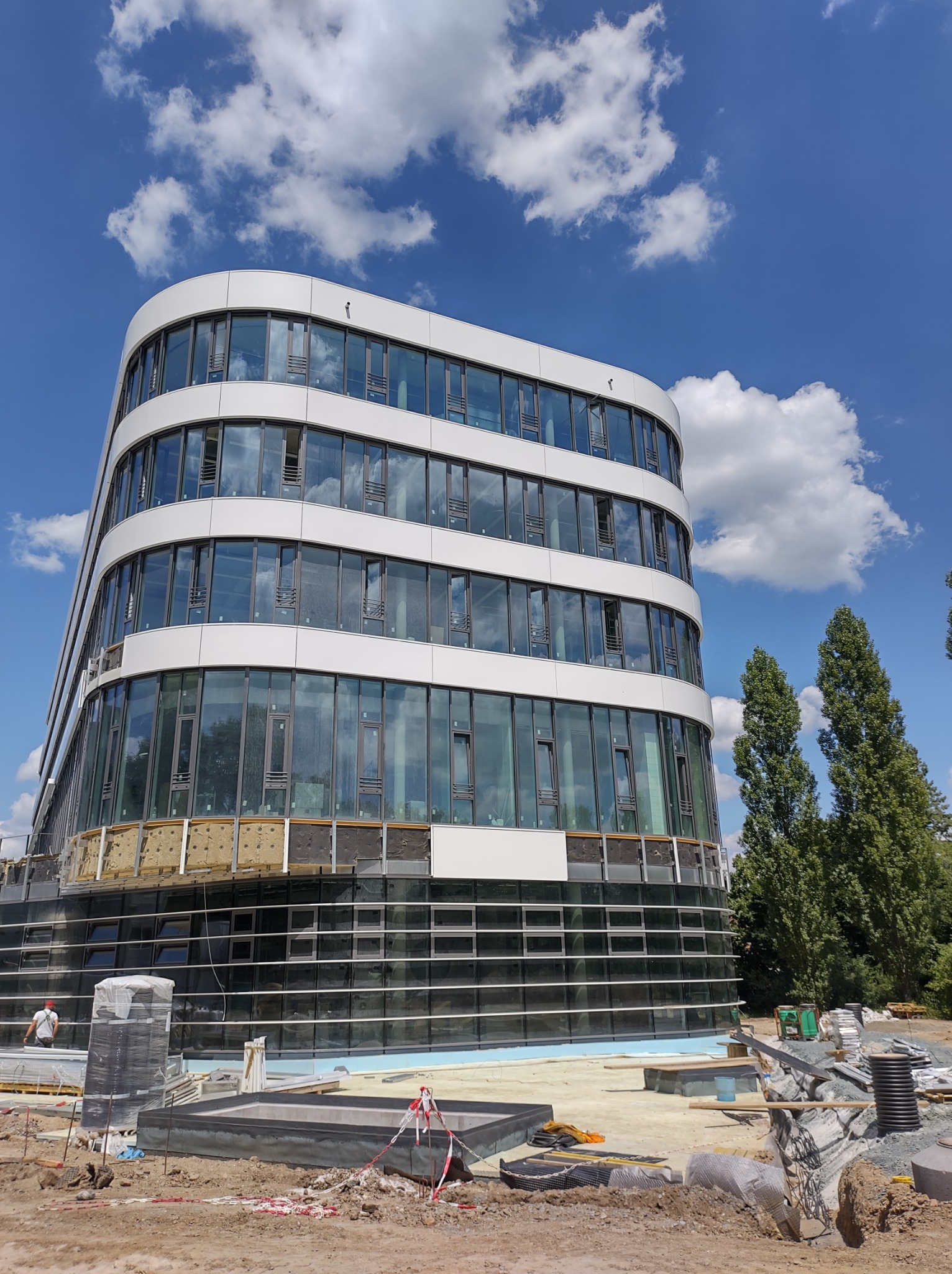 Nowoczesny, zaokrąglony budynek biurowy z szklaną fasadą w trakcie budowy, widoczne materiały izolacyjne na dolnych kondygnacjach i robotnik w oddali, błękitne niebo z chmurami w tle.