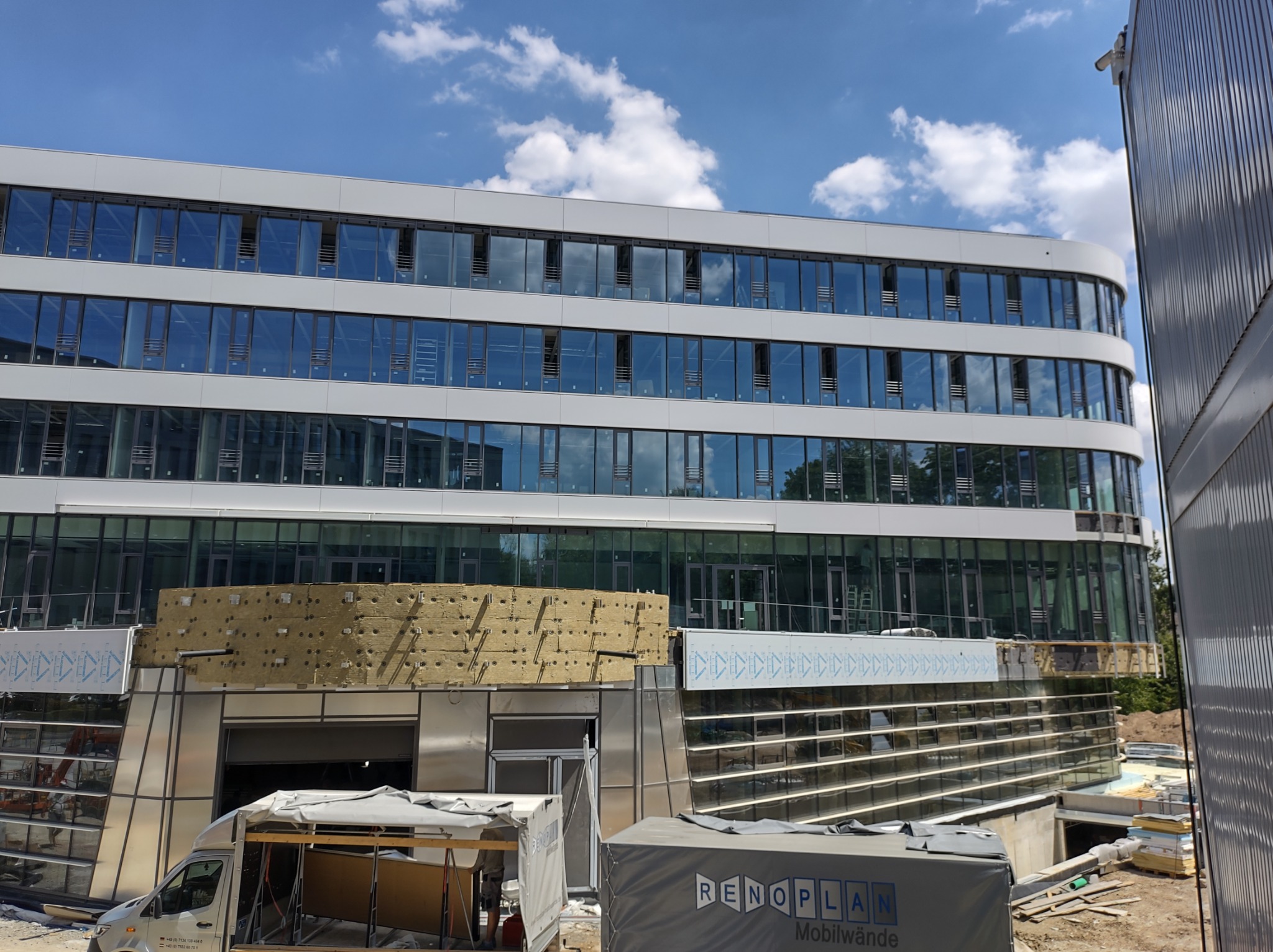 Wielopiętrowy budynek biurowy z częściowo zamontowaną elewacją, widoczne płyty izolacyjne i elementy konstrukcyjne, na pierwszym planie samochód dostawczy i kontener z napisem RENOPLAN.