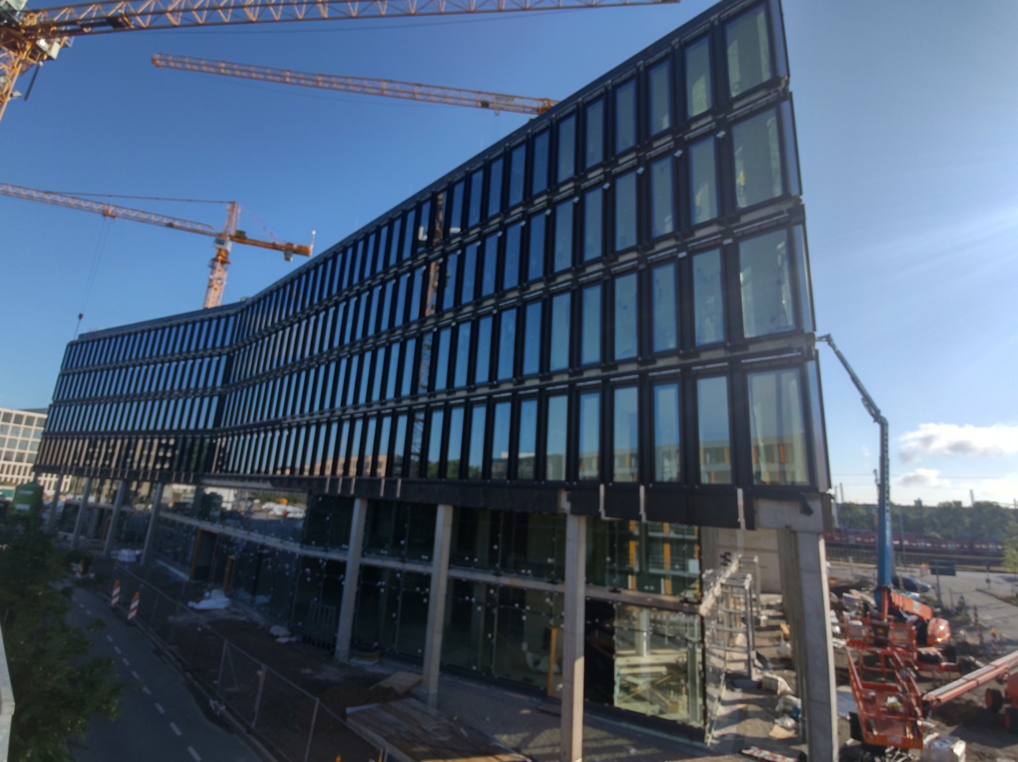 Wysoki budynek biurowy w trakcie budowy z widocznymi dźwigami i podnośnikiem koszowym, nowoczesna fasada z licznymi oknami odbijającymi niebo.