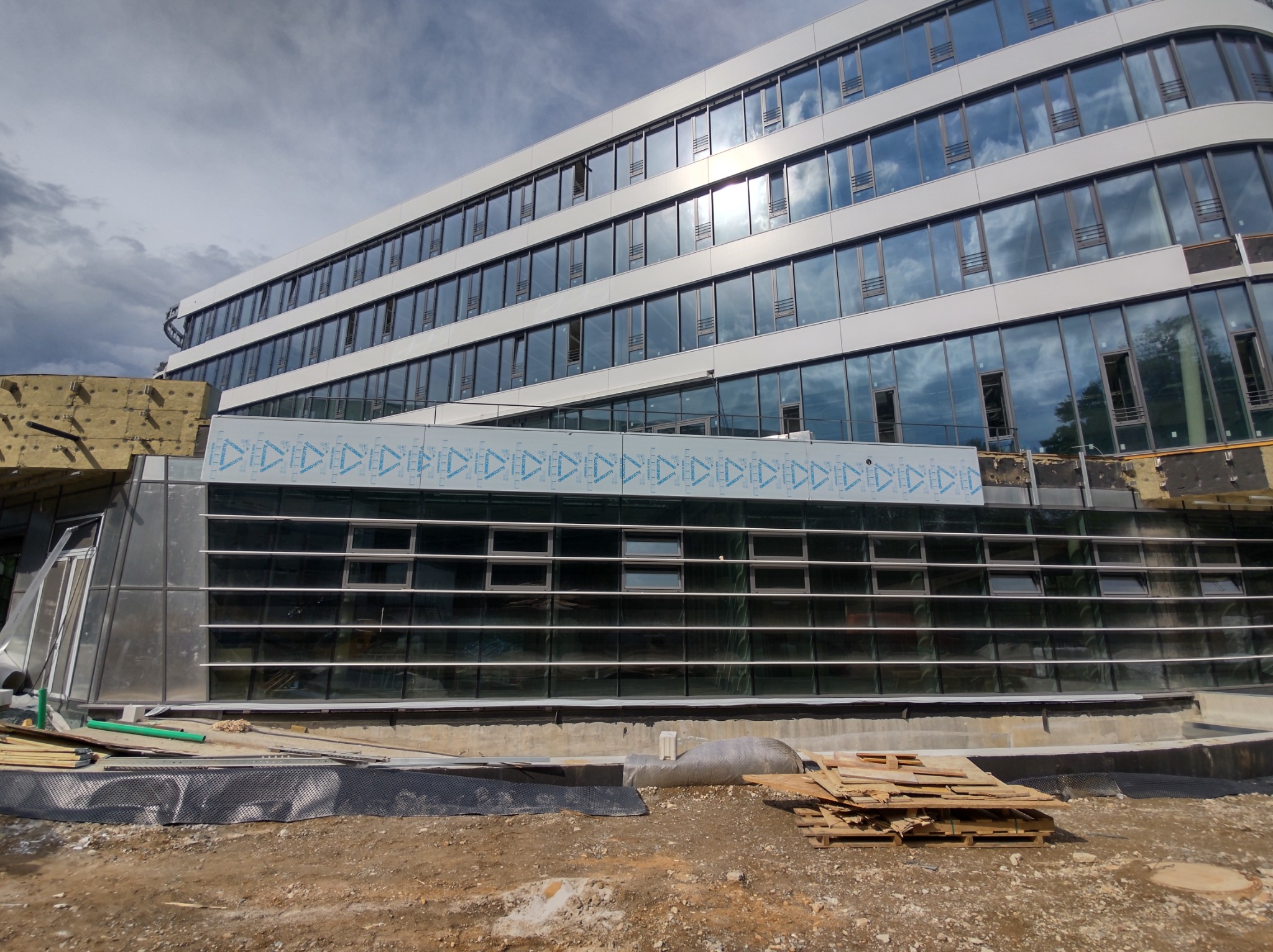 Wielopiętrowy budynek biurowy w trakcie montażu elewacji, widoczne płyty elewacyjne z folią ochronną, ocieplenie w górnej części i materiały budowlane na ziemi.