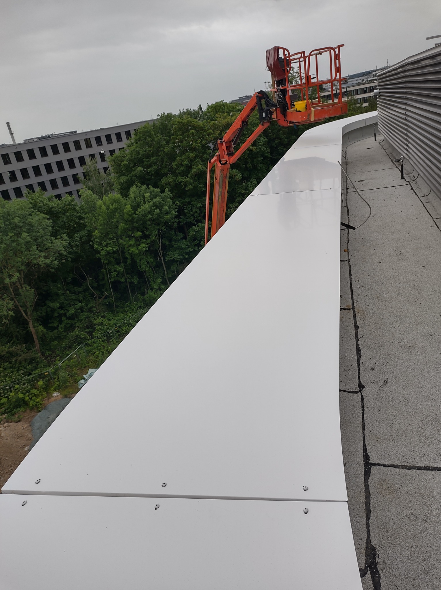 Pomarańczowy podnośnik koszowy na dachu budynku z zamontowanymi białymi panelami elewacyjnymi, widoczne drzewa i budynek w tle.