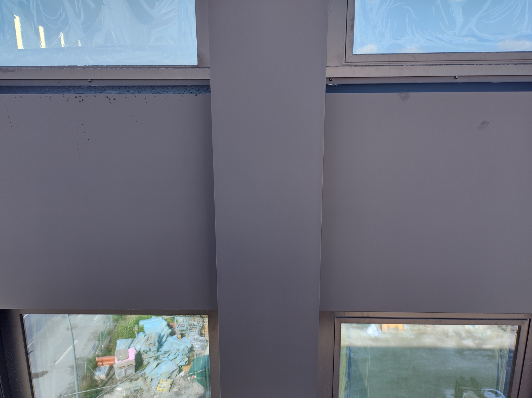 Szare panele elewacyjne z widocznymi oknami, zabezpieczonymi folią, oraz widokiem na plac budowy z elementami rusztowań i materiałów budowlanych.