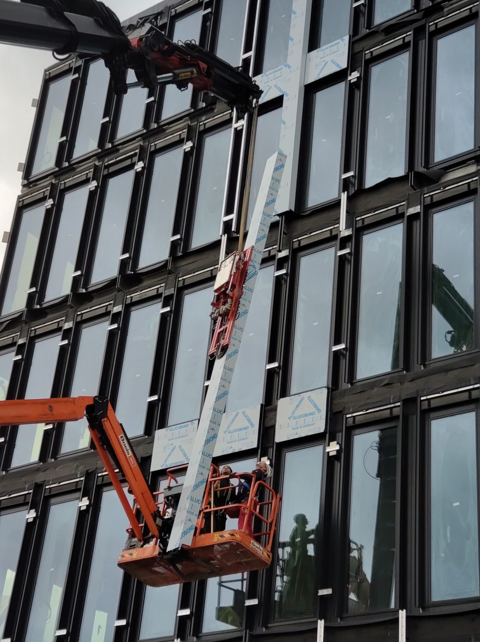 Montaż paneli elewacyjnych Alucobond na fasadzie budynku biurowego przy użyciu podnośnika koszowego i żurawia. Widoczne elementy mocujące i okna. Prace w toku.