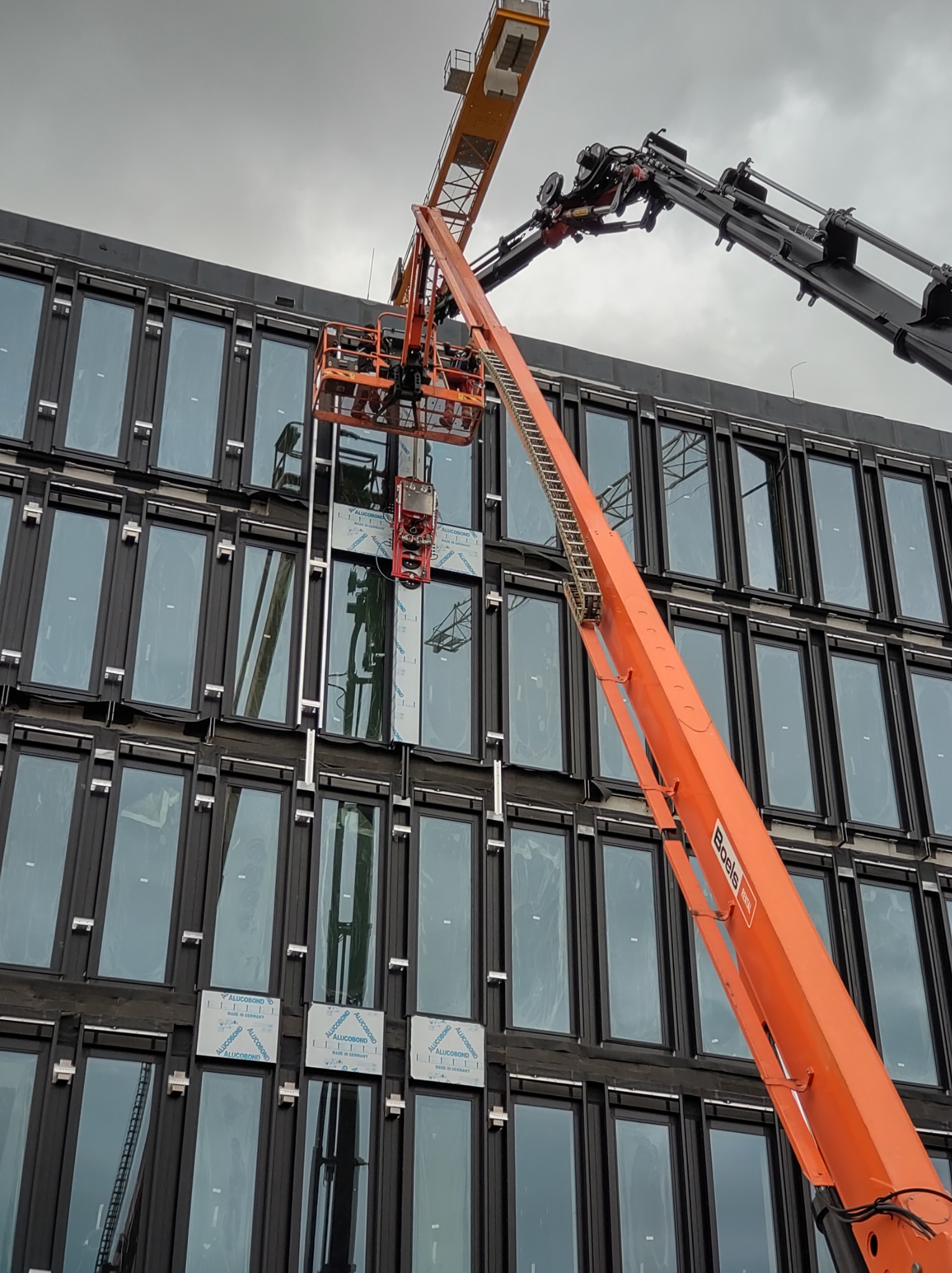 Montaż paneli Alucobond na elewacji budynku biurowego, widoczny pomarańczowy podnośnik koszowy Boels Rental i dźwig w tle, prace na wysokościach.