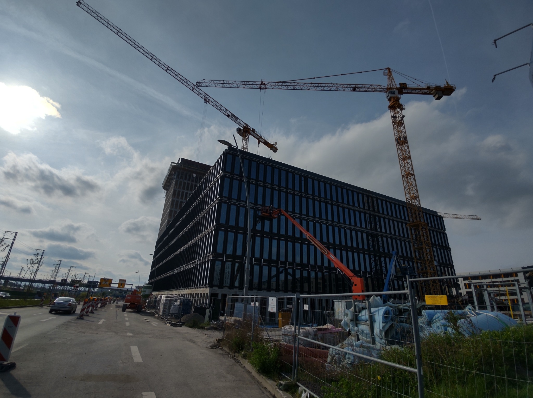 Budynek biurowy w trakcie budowy z ciemną szklaną fasadą, widoczne dwa żurawie budowlane, podnośnik koszowy, ogrodzenie placu budowy oraz ruch uliczny w tle.