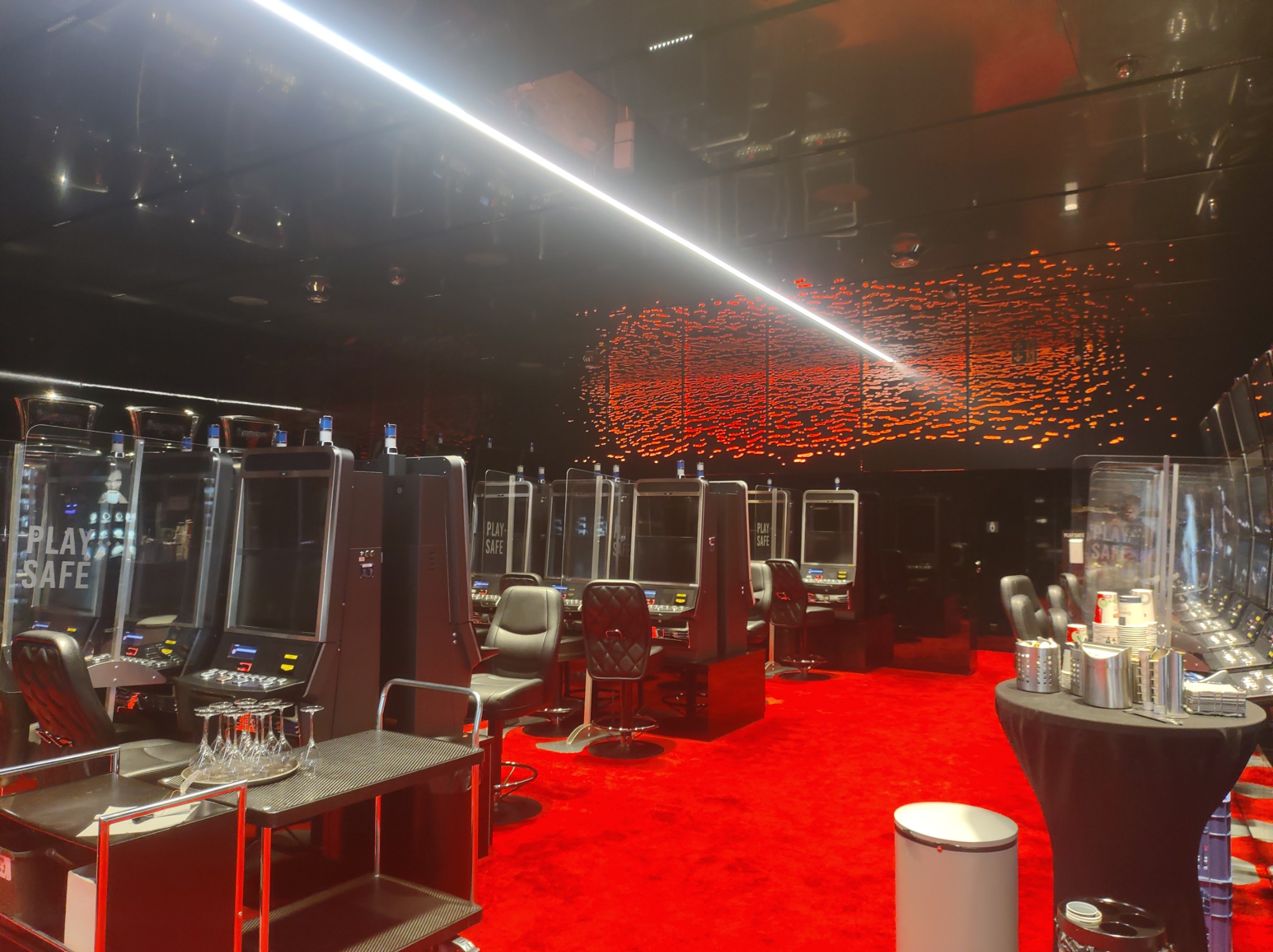 Wnętrze kasyna z rzędem automatów do gier oddzielonych pleksiglasem, czerwona wykładzina, czarne ściany z dekoracją imitującą płomienie, stolik z szklankami i wózkiem barowym.