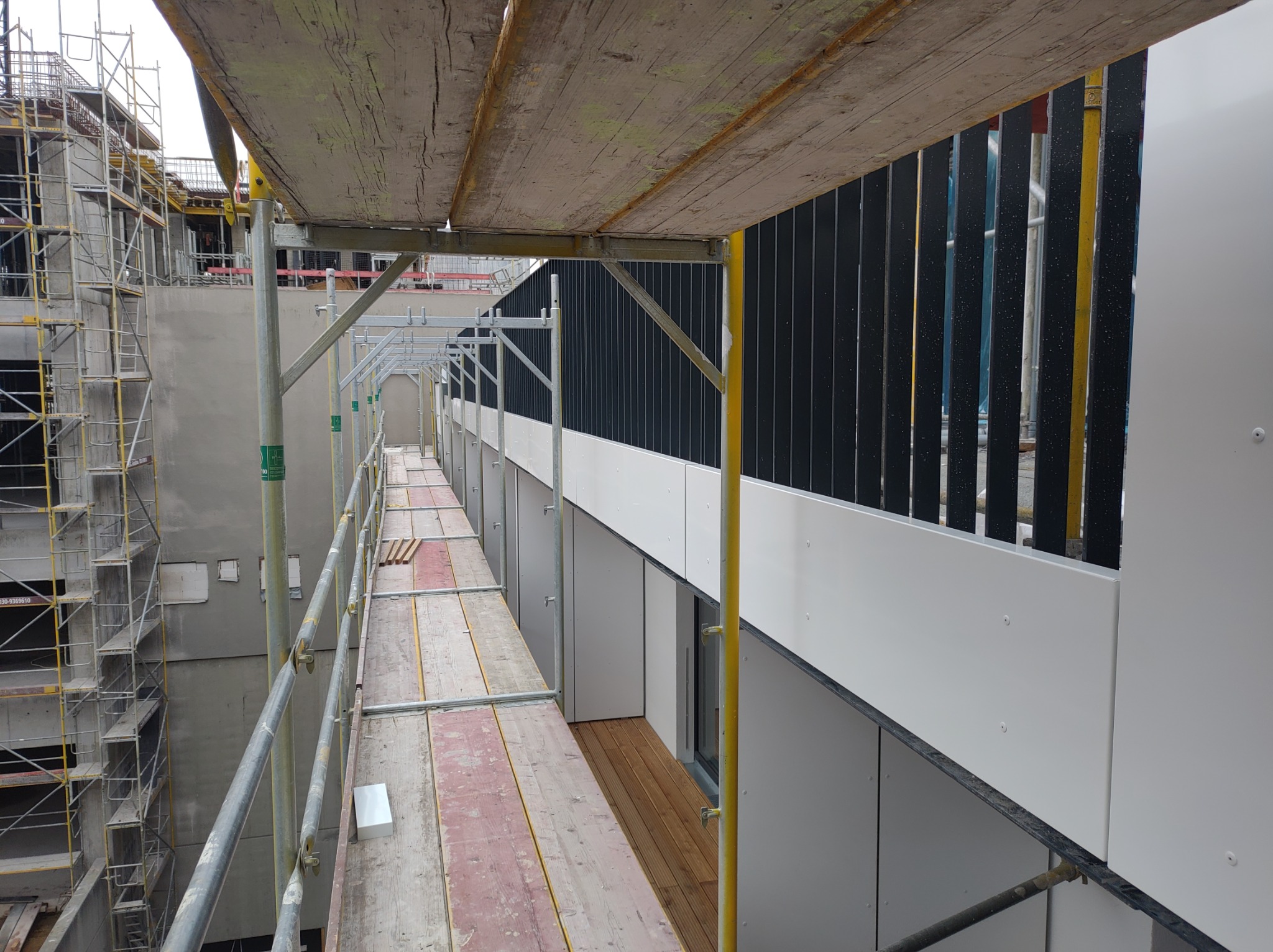 Widok rusztowania z drewnianymi podestami wzdłuż ściany budynku z zamontowanymi białymi panelami elewacyjnymi i czarnymi pionowymi elementami dekoracyjnymi. W tle widoczne kolejne rusztowania...